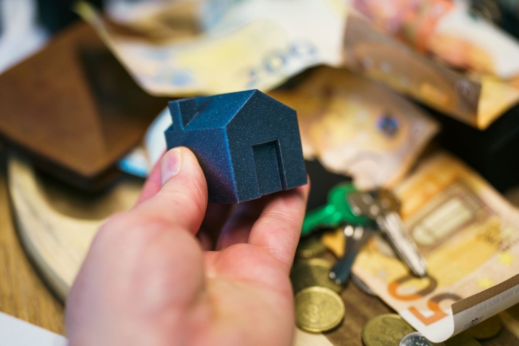 Miniature house model held in hand next to cash, keys, and a wallet, symbolizing choosing between a mortgage and a cash property purchase