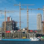 Tower cranes over a residential building site on a waterfront, with a cargo barge moored in the foreground