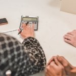 Adviser using a calculator beside a purchase real estate form while two buyers wait with hands folded