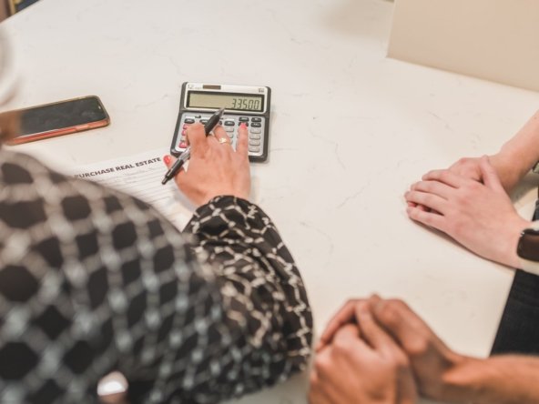 Adviser using a calculator beside a purchase real estate form while two buyers wait with hands folded