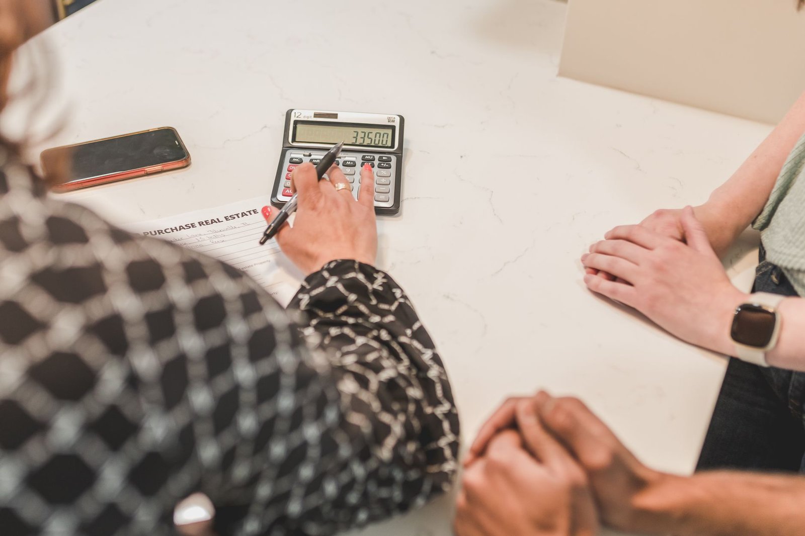 Adviser using a calculator beside a purchase real estate form while two buyers wait with hands folded