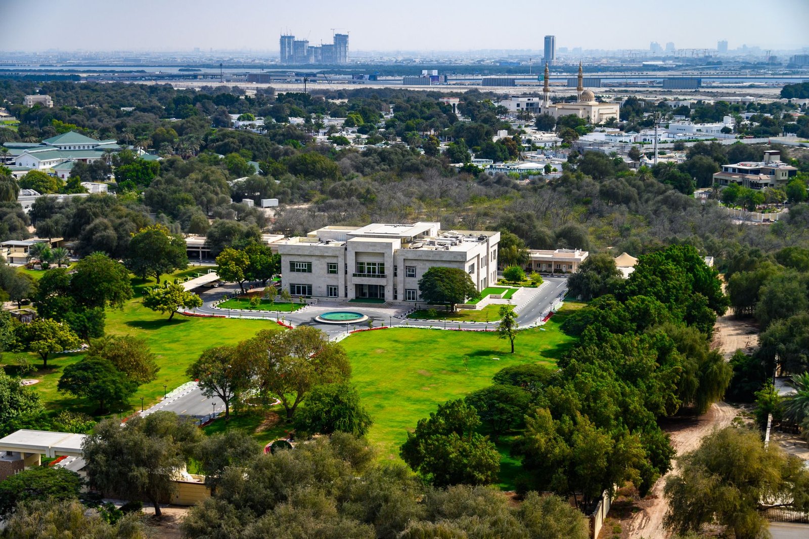 Aerial view of a landscaped Dubai estate with green lawns, mature trees and distant city towers on the horizon