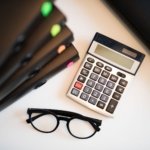 Overhead view of a calculator and reading glasses beside stacked document folders on an office desk, illustrating preparing a tax return for overseas property