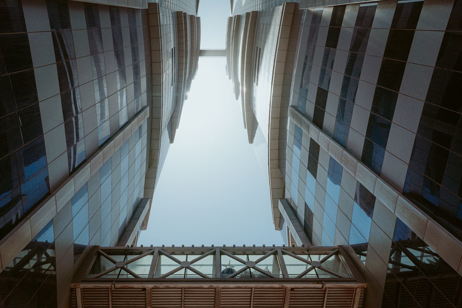 Looking up between two stone and glass office towers joined by a sky bridge under a clear sky